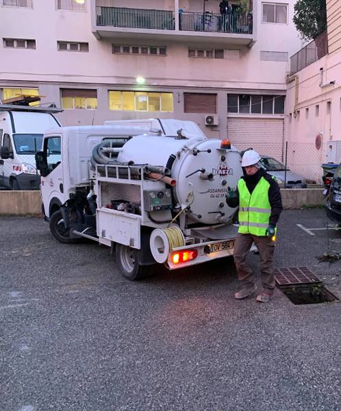 Un ouvrier casqué en gilet jaune fluorescent se tient à côté d'un camion hydrocureur garé sur un parking, près d'une grille d'évacuation.