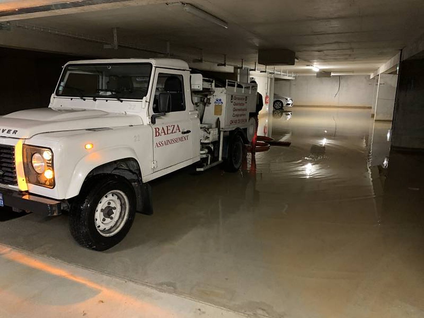 Un camion blanc de l'entreprise Baeza Assainissement est garé dans un parking souterrain dont le sol est recouvert d'eau boueuse.
