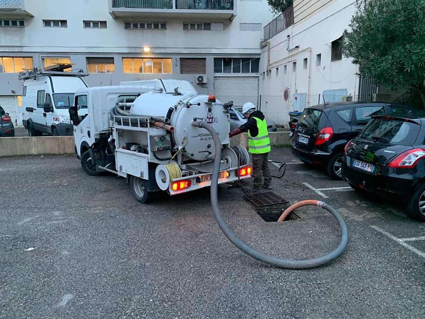 Un technicien en gilet jaune fluo utilise un camion hydrocureur et un gros tuyau pour intervenir sur une bouche d'égout dans un parking.