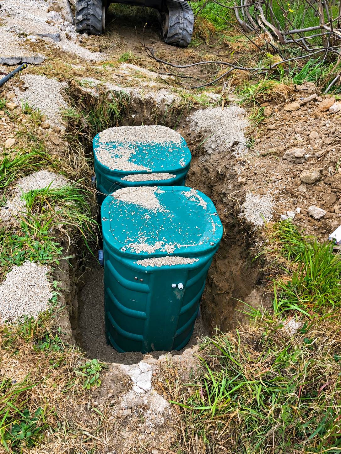 Deux cuves vertes recouvertes de gravier sont installées dans une tranchée creusée dans un terrain herbeux, avec un engin de chantier visible en haut de l'image.