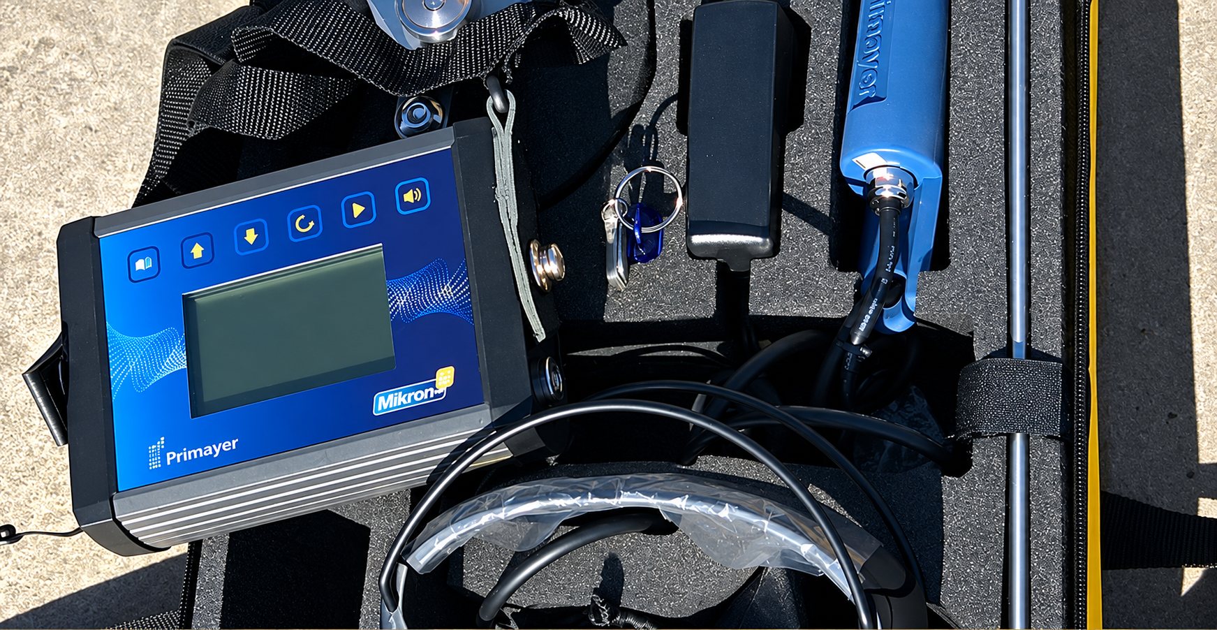 Un appareil électronique Primayer Mikron avec écran et boutons, posé dans une mallette en mousse à côté d'un casque audio, d'une sonde bleue et d'une tige métallique.
