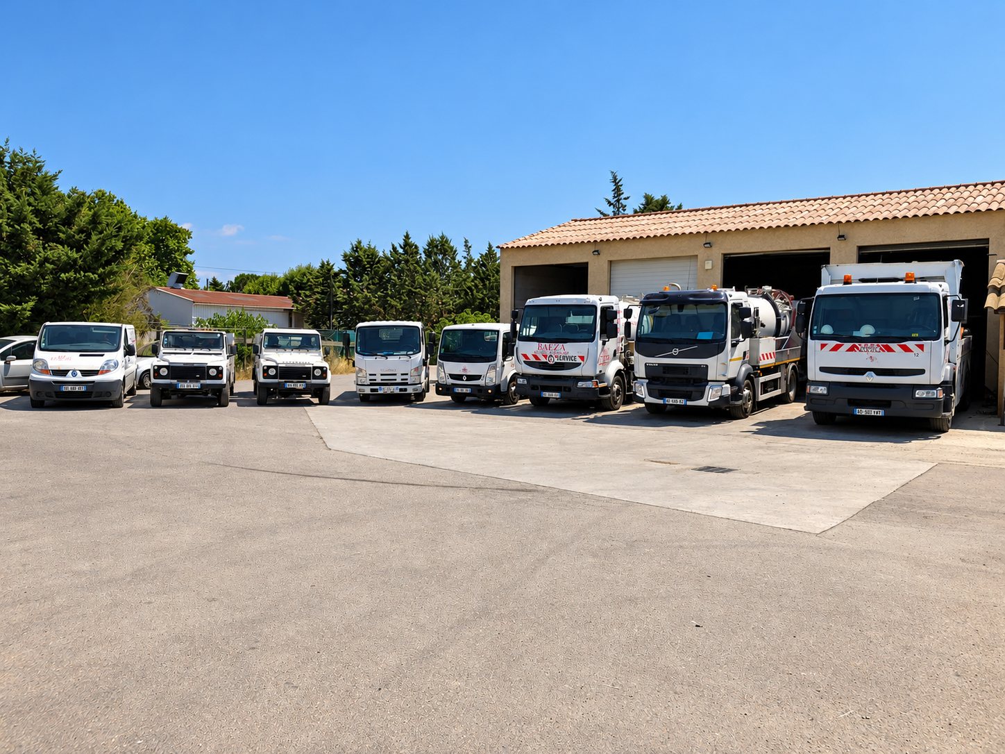 Une flotte de camions blancs, dont des camions hydrocureurs, de fourgonnettes et de 4x4, est alignée sur une cour devant un entrepôt à toit de tuiles.