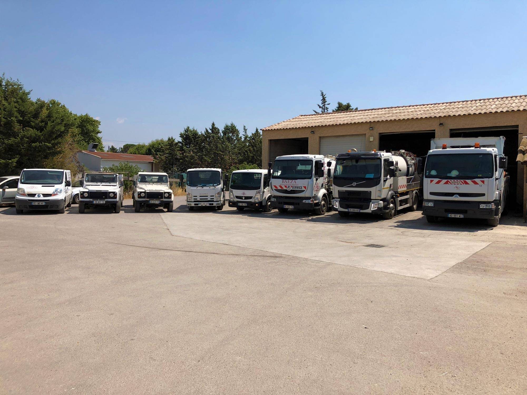 Une flotte de camions blancs, dont des camions hydrocureurs, de fourgonnettes et de 4x4, garée devant un hangar au toit de tuiles sous un ciel bleu.