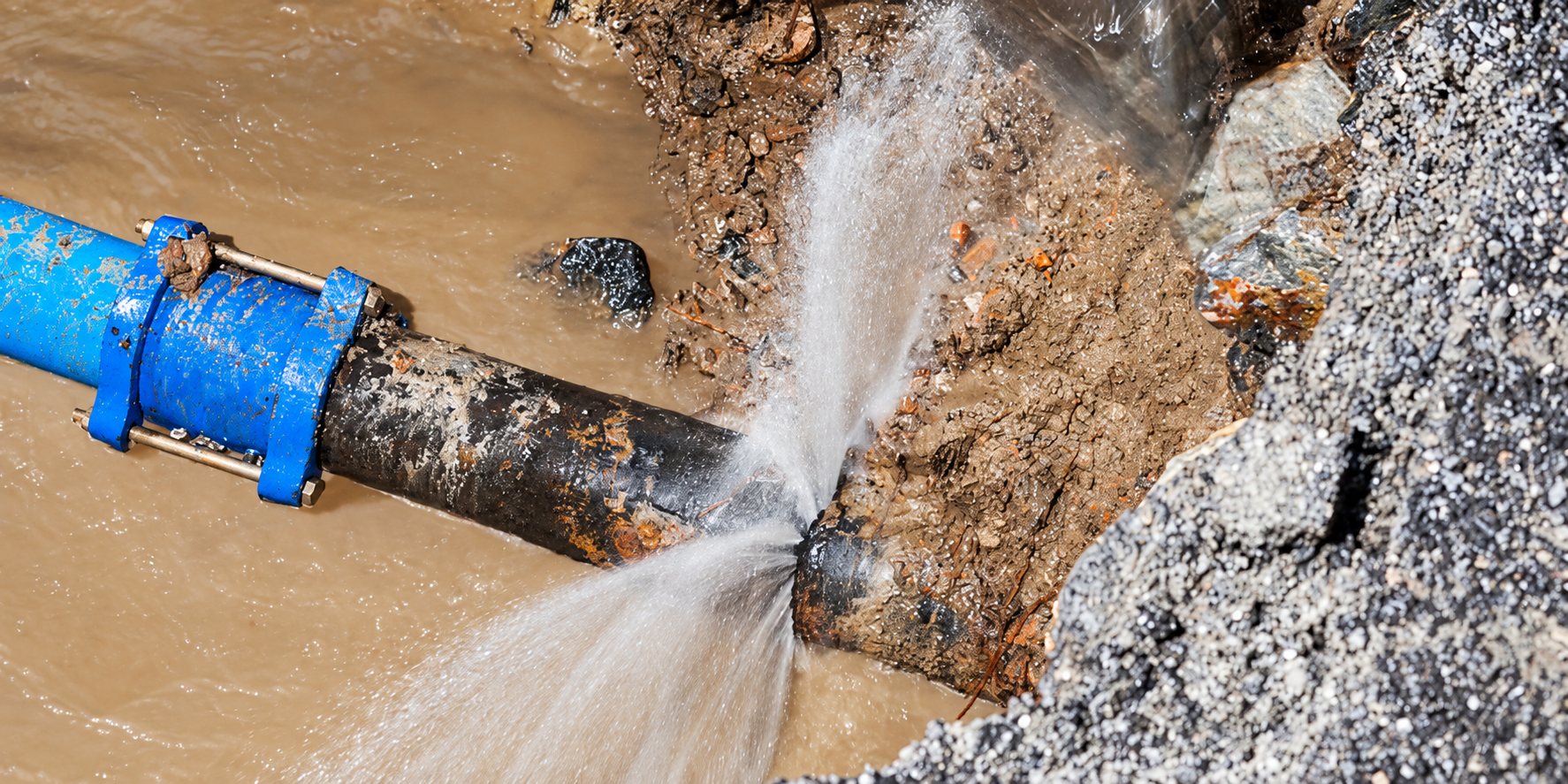 Une canalisation rouillée enterrée dans une tranchée boueuse laisse jaillir un jet d'eau près d'un collier de réparation bleu.
