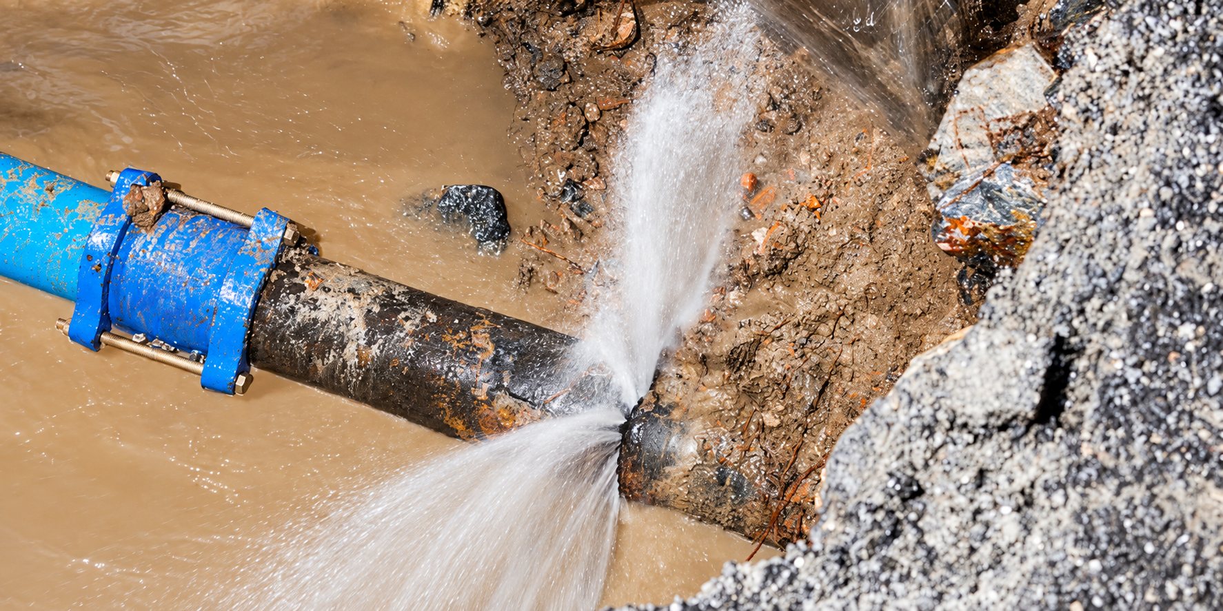 Une conduite métallique rouillée avec un collier de réparation bleu laisse jaillir un jet d'eau dans une tranchée remplie de boue et de gravats.