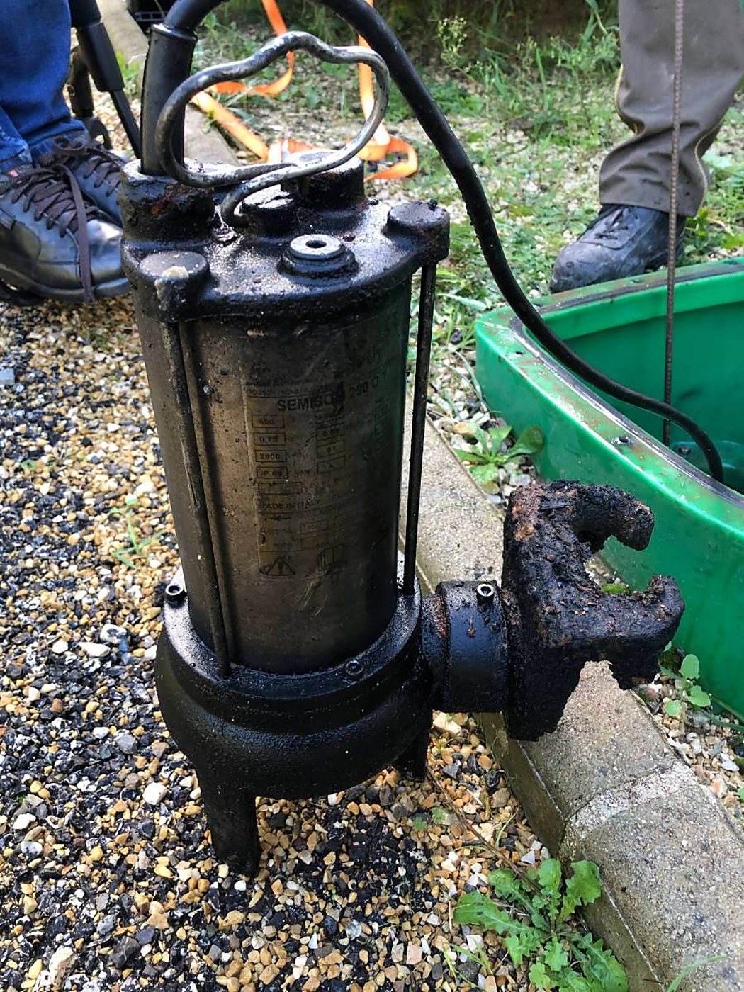 Une pompe de relevage immergée recouverte de dépôts noirs est posée sur du gravier, à côté d'une bordure en béton et d'une cuve verte.