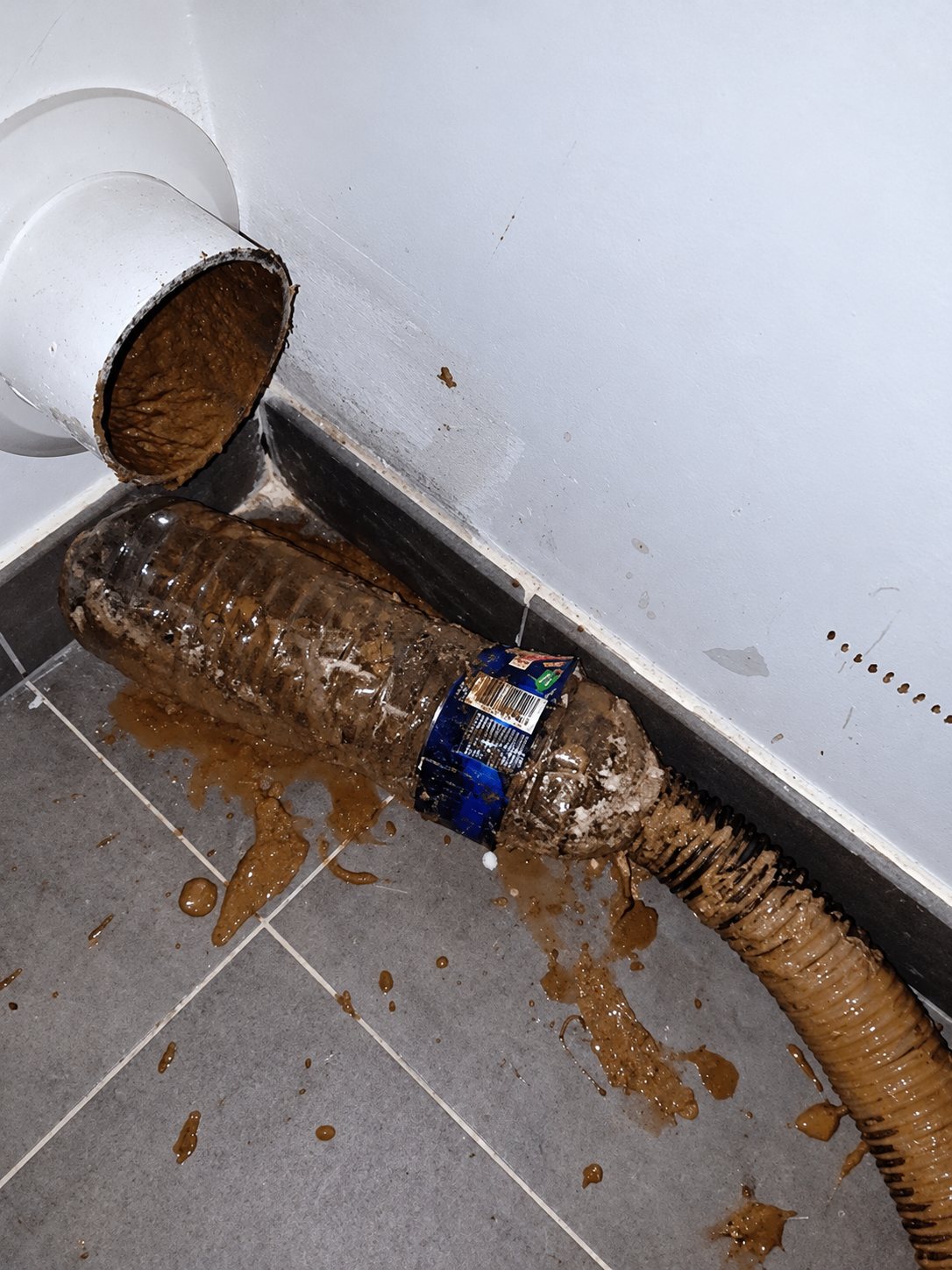 Un tuyau d'évacuation blanc fixé au mur déverse de la boue brune dans une bouteille en plastique et un flexible posés sur un carrelage gris sali.